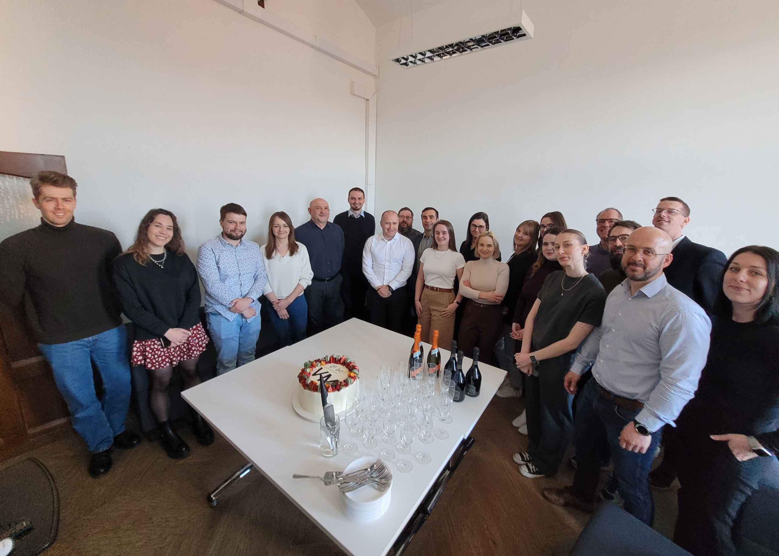 A group of colleagues gathered around a table in an office to celebrate a special occasion. The table is set with an anniversary cake, champagne bottles, glasses, and serving utensils as the team poses for a group photo.