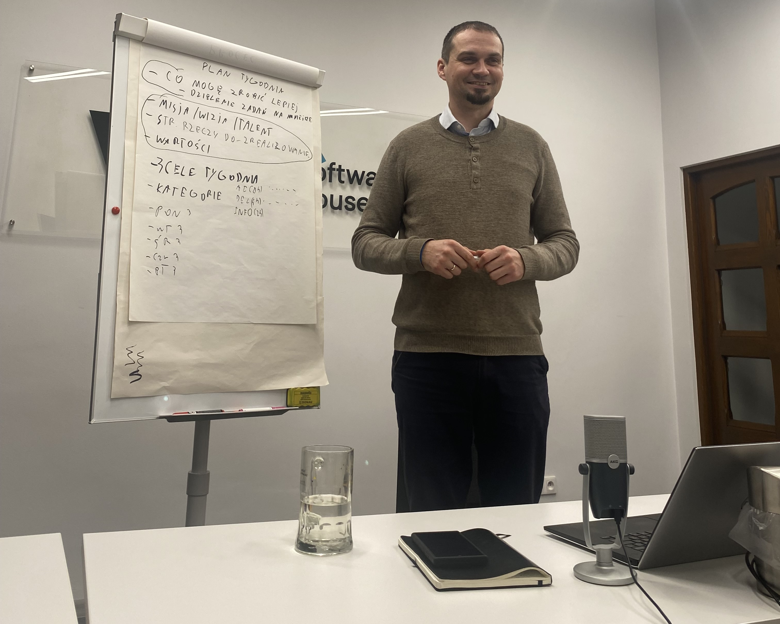 Łukasz Borzęcki delivering a team workshop in a meeting room, standing next to a flip chart with planning notes and discussing goal setting, personal development, and weekly planning with participants.