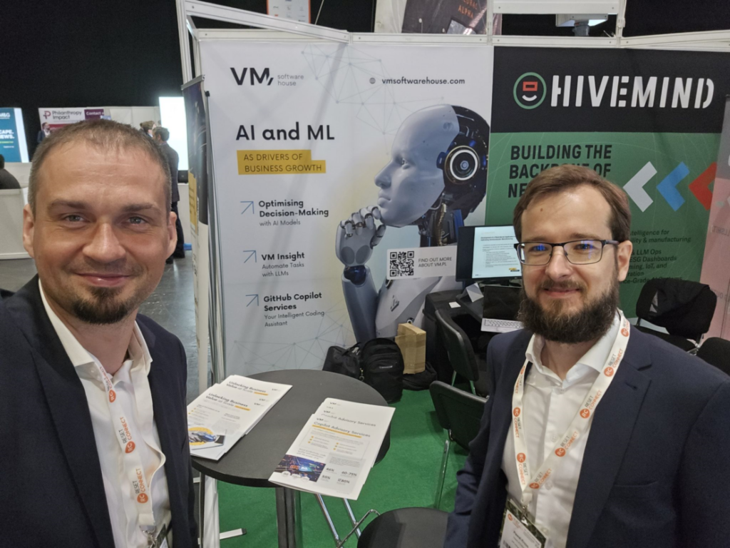 Two VM.pl representatives standing at the company's exhibition booth during a technology conference, with banners showcasing AI, machine learning, decision support, GitHub Copilot services, and business automation solutions.