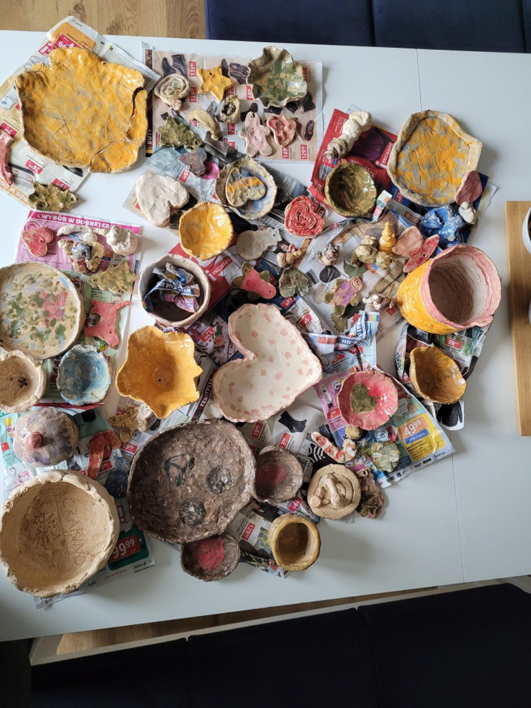 Handmade and colorfully decorated ceramic creations made by children during a St. Nicholas Day workshop for employees' families. The table is filled with bowls, ornaments, figurines, and painted clay crafts left to dry.