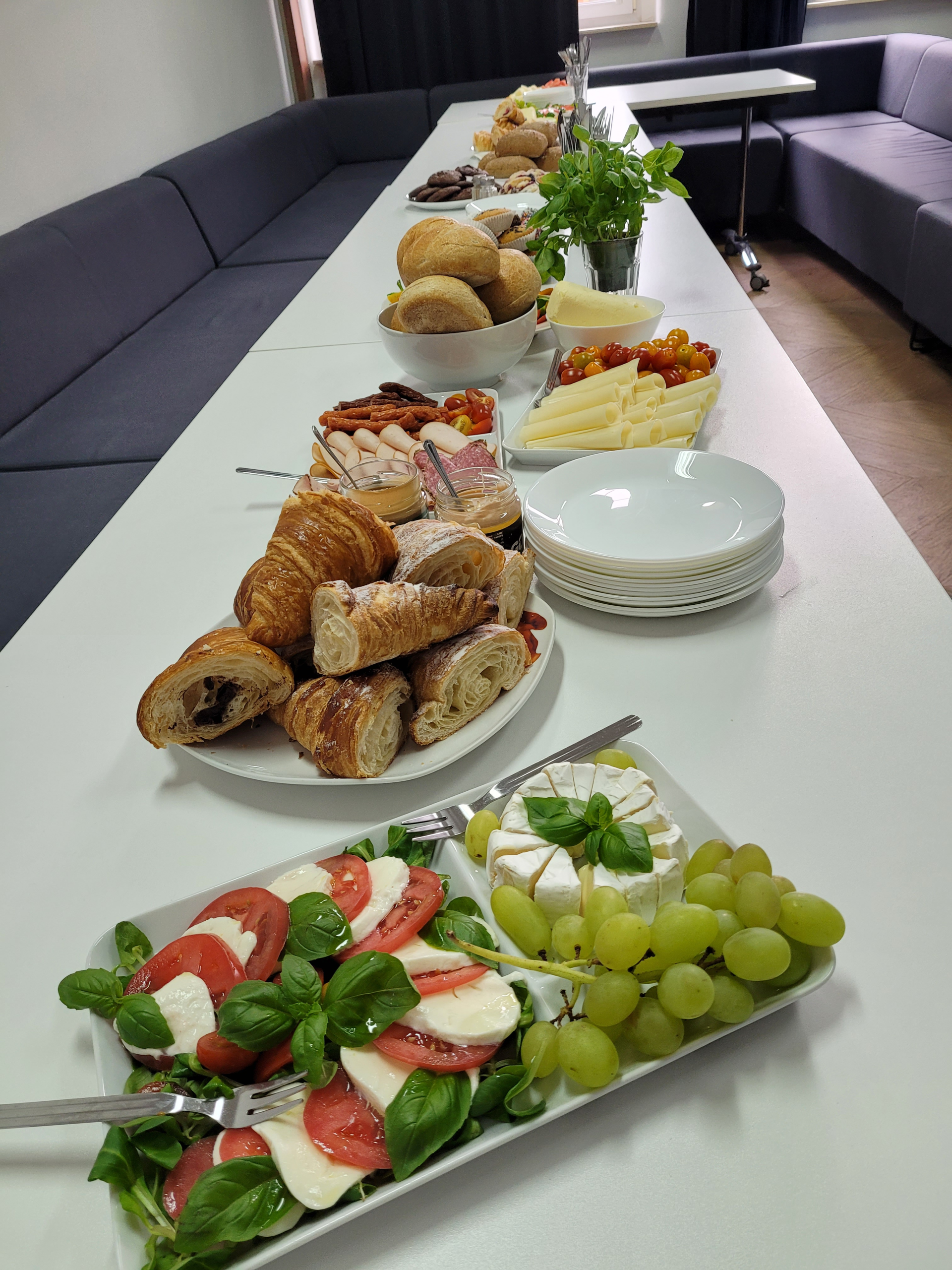 A breakfast buffet prepared for Men's Day at the office, featuring croissants, fresh bread, cheeses, cold cuts, vegetables, fruit, soft cheese, and plates ready for employees to enjoy together.