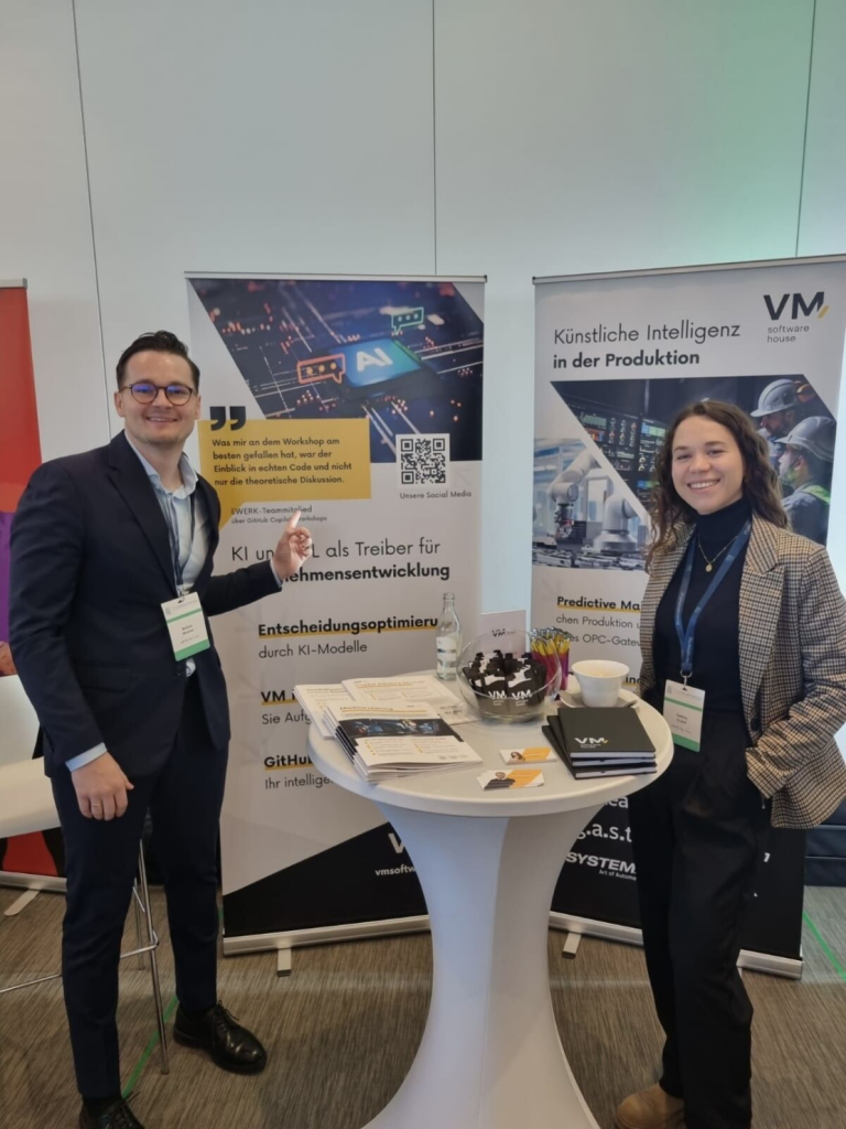 Zaklina Kozioł and Mariusz Mrowiec representing VM.pl at a technology conference, standing beside the company's exhibition booth showcasing artificial intelligence and machine learning solutions for manufacturing and business process optimization.