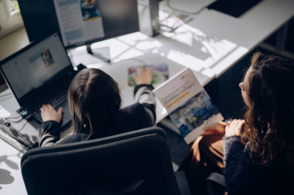 Photo of two VM.PL employees reviewing project materials at a workstation. One team member works on a laptop while the other discusses a printed document in a collaborative office environment.