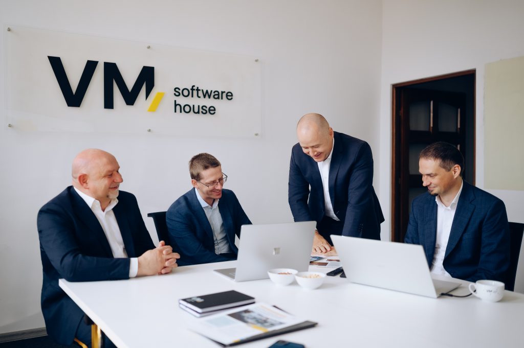 Photo of the VM.PL management board during a business meeting at the company's headquarters. Board members are discussing projects around a conference table with laptops.